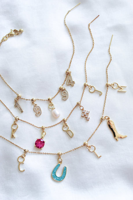 Kinsey Designs' Charm Bracelet Dainty Chain is styled with gold necklaces featuring number, letter, and charm pendants on white fabric for a chic layered look.
