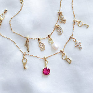 Kinsey Designs’ gold necklaces with letter and number charms, a pink gem, pearl, rhinestone details, and a Silver Birthstone Grip Charm are displayed on a white fabric background.