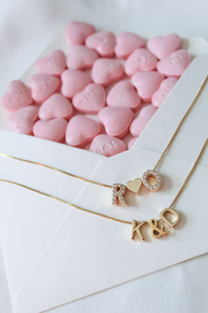 Gold filled necklaces with "R♥D" and "K&D" letter pendants rest on an envelope of pink heart candies, accented by the Ampersand Spacer Slide from Kinsey Designs’ Charm Bar collection.