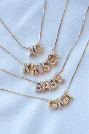 Four gold necklaces from Kinsey Designs, each with a LUX Letter Slide Charm—XO, KINSEY, BABE, and GIGI—rest on white fabric, featuring sparkling crystal-studded pendants for a personalized touch.