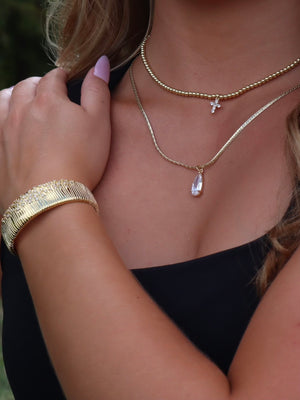 A woman in a black top shows manicured nails and gold bracelets, highlighting the Kinsey Designs Charlotte Drop Necklace—a gold-filled teardrop piece with a sparkling cubic zirconia pendant for an elegant touch.