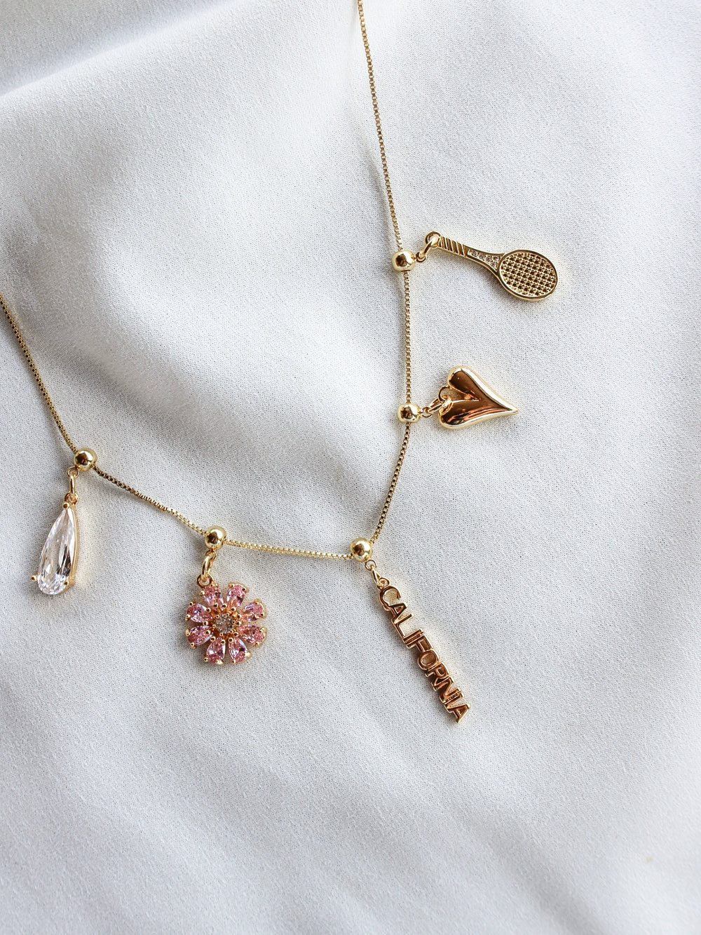 Texas Grip Charm necklace by Kinsey Designs features a tennis racket, heart, teardrop gem, pink flower, and “CALIFORNIA” text on fabric—ideal for a playful touch to your jewelry collection.