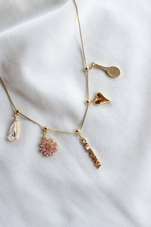 Texas Grip Charm necklace by Kinsey Designs features a tennis racket, heart, teardrop gem, pink flower, and “CALIFORNIA” text on fabric—ideal for a playful touch to your jewelry collection.