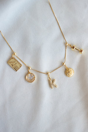 The Kinsey Designs Smiley Grip Charm necklace features five unique charms—a smiley, mother-of-pearl, moon, letter K, and a tiny dumbbell—elegantly displayed on white fabric.
