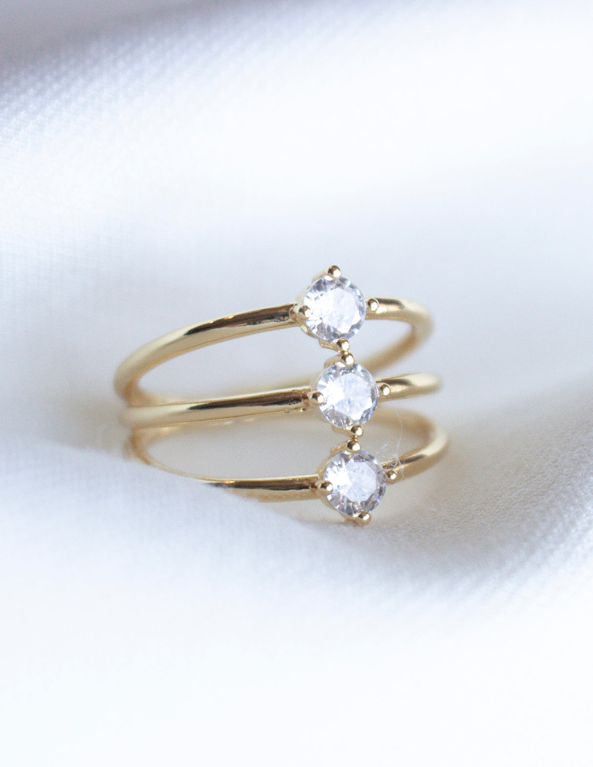 The Indy Ring by Kinsey Designs Archives features three round clear cubic zirconia stones set in a row on a gold filled band, shown against a white fabric background.