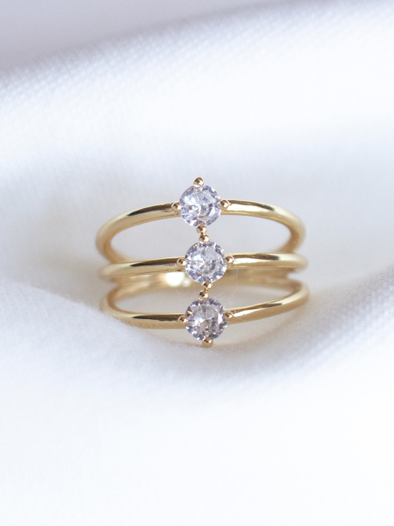 The Indy Ring by Kinsey Designs Archives is a gold filled ring with three bands, each set with a round cubic zirconia, shown on a white fabric background.