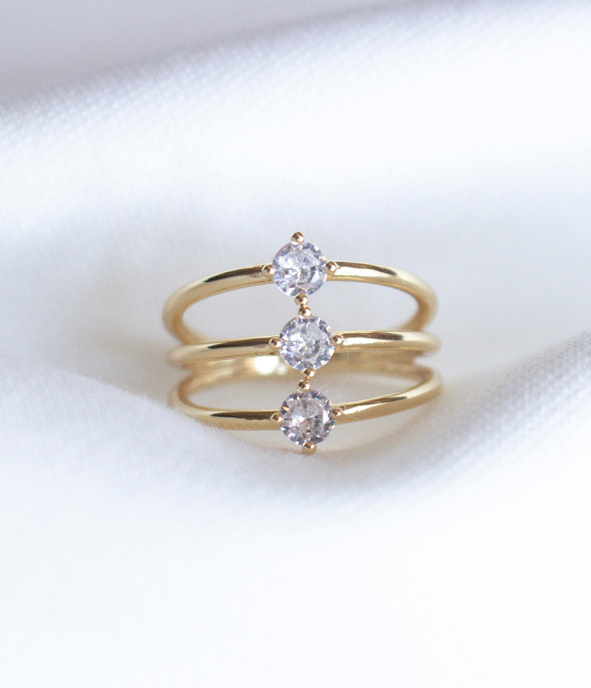The Indy Ring by Kinsey Designs Archives is a gold filled ring with three bands, each set with a round cubic zirconia, shown on a white fabric background.