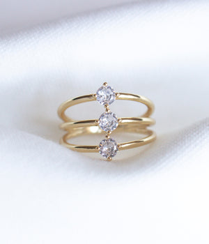 The Indy Ring by Kinsey Designs Archives is a gold filled ring with three bands, each set with a round cubic zirconia, shown on a white fabric background.
