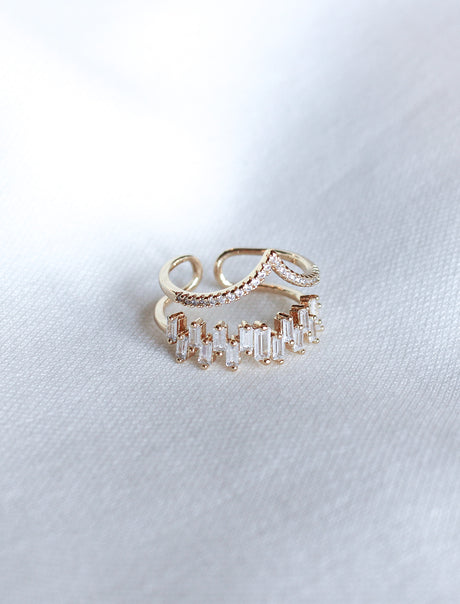 The Ines Ring by Kinsey Designs is a gold-plated double-band ring adorned with sparkling cubic zirconia, shown on a white textured fabric background.