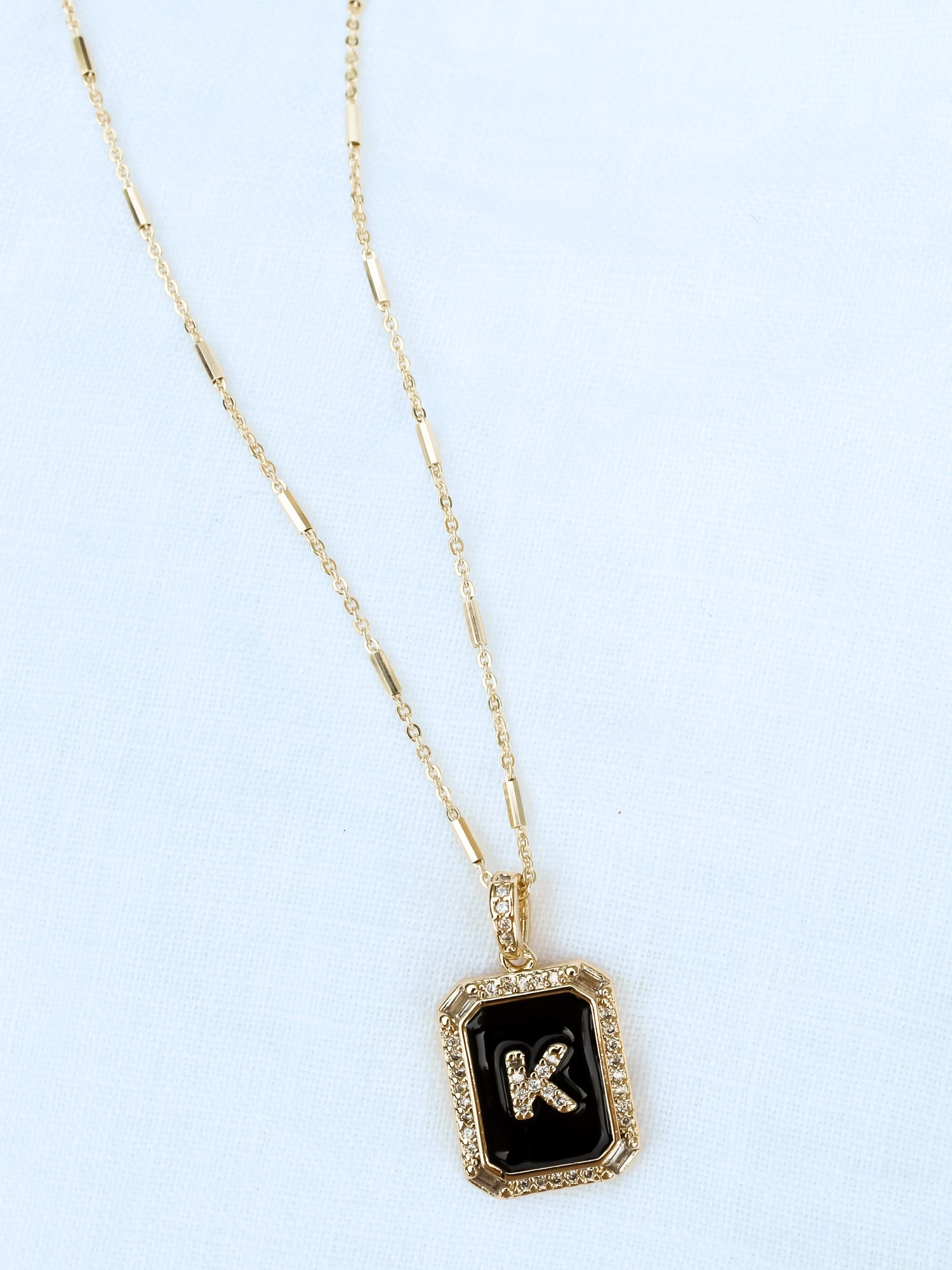 The Initial Tile Necklace Ink Letter K by Kinsey Designs Archives features a gold chain and a black pendant with the letter "K" on a light textured background.