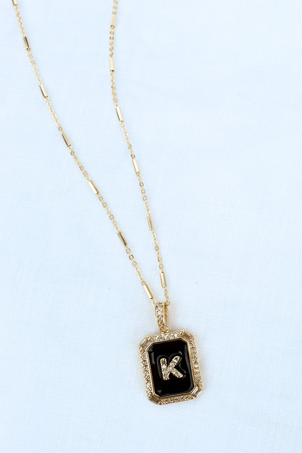The Initial Tile Necklace Ink Letter K by Kinsey Designs Archives features a gold chain and a black pendant with the letter "K" on a light textured background.
