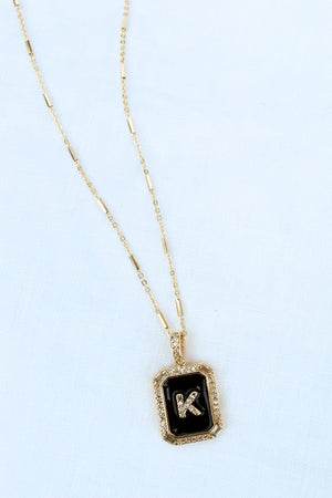 The Initial Tile Necklace Ink Letter K by Kinsey Designs Archives features a gold chain and a black pendant with the letter "K" on a light textured background.