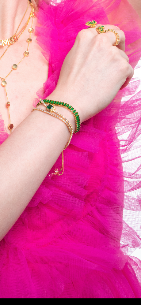 A woman in a pink ruffled dress wears gold jewelry, including Kinsey Designs Archives’ Ivy Bracelet, along with green bracelets and gold rings set with green stones.