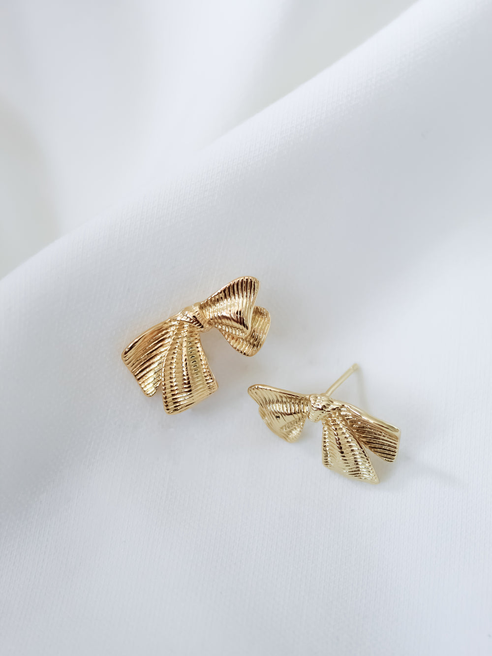 Gold-tone Izzy Bow Studs by Kinsey Designs, displayed on white fabric, add a touch of elegance to any outfit.
