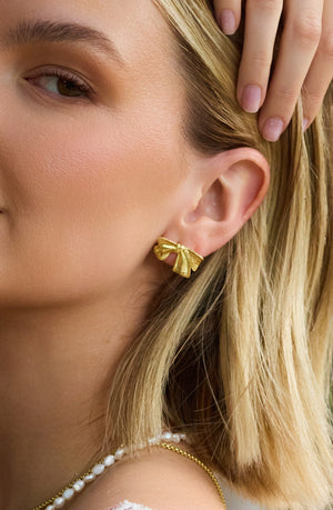 Close-up of a woman wearing Kinsey Designs’ Izzy Bow Studs, gold-tone bow earrings paired with a pearl necklace, her hand gently resting by her ear.