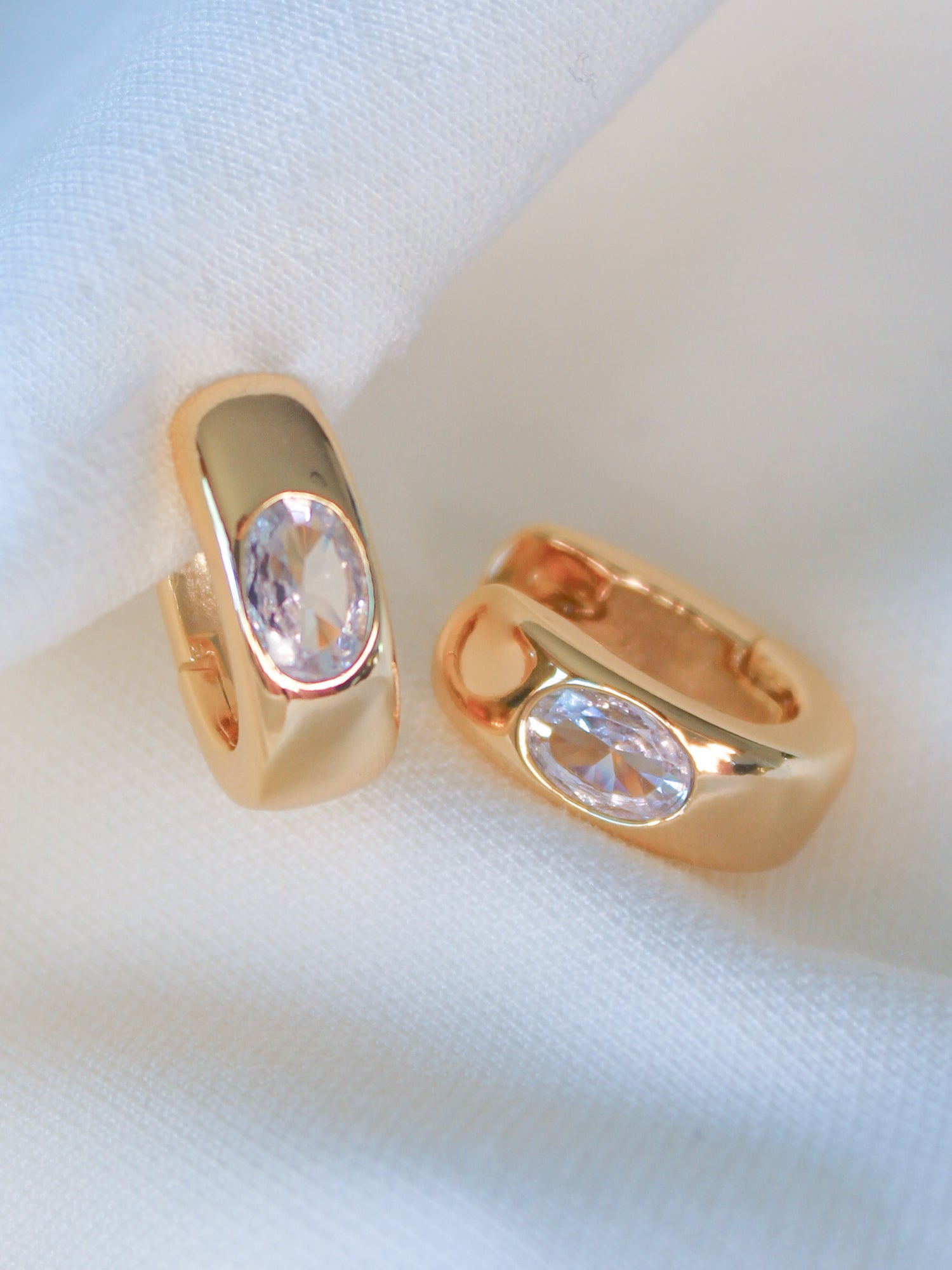 The Jack Huggie by Kinsey Designs features gold-filled earrings with oval clear gemstones, displayed on soft white fabric. These sample sale pieces add an elegant touch to any look.