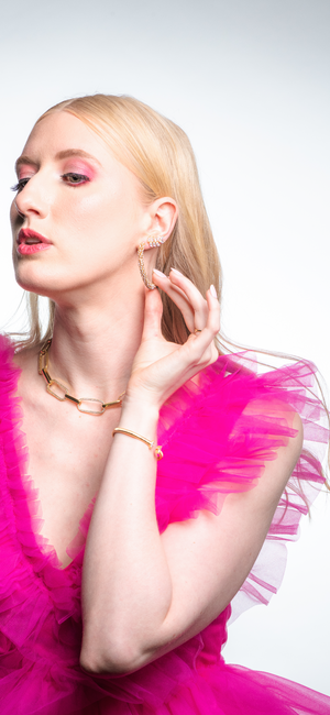 A woman in a bright pink ruffled dress touches her gold earring and wears the Kinsey Designs Jameson Necklace, paired with bold jewelry and pink makeup.
