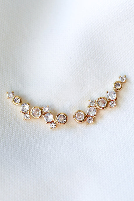 The Jax Climber by Kinsey Designs Archives features gold-plated earrings with sparkling cubic zirconia set in a curved design, displayed on a white fabric background.