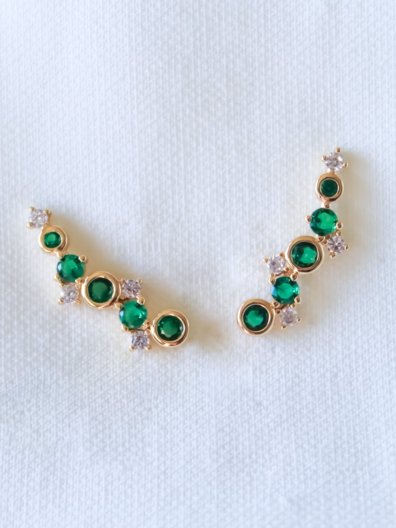 The Jax Climber-Emerald by Kinsey Designs Archives are gold plated earrings, 1" wide, featuring green and white cubic zirconia in a curved design on a white textured background.