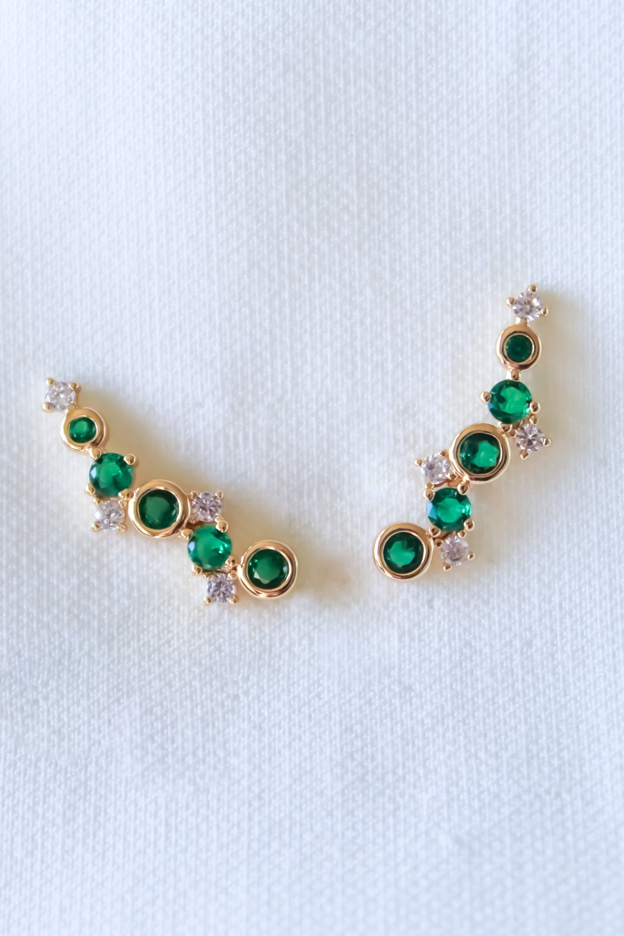 The Jax Climber-Emerald by Kinsey Designs Archives are gold plated earrings, 1" wide, featuring green and white cubic zirconia in a curved design on a white textured background.