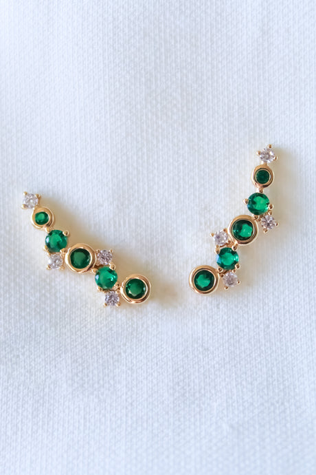 The Jax Climber-Emerald by Kinsey Designs Archives are gold plated earrings, 1" wide, featuring green and white cubic zirconia in a curved design on a white textured background.