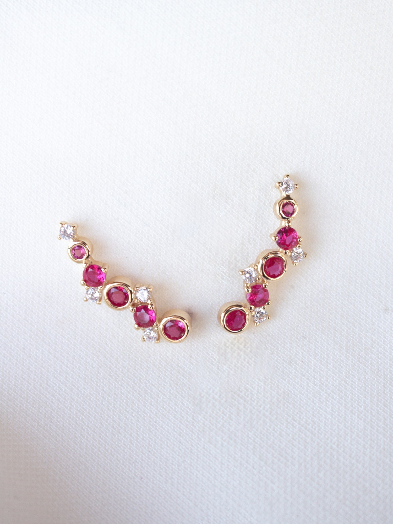 The Jax Climber Fuchsia earrings by Kinsey Designs feature gold filled curves adorned with vibrant fuchsia gemstones and sparkling cubic zirconia, beautifully displayed on a white fabric background.
