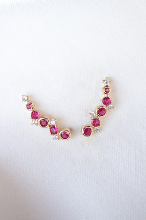 The Jax Climber Fuchsia earrings by Kinsey Designs feature gold filled curves adorned with vibrant fuchsia gemstones and sparkling cubic zirconia, beautifully displayed on a white fabric background.
