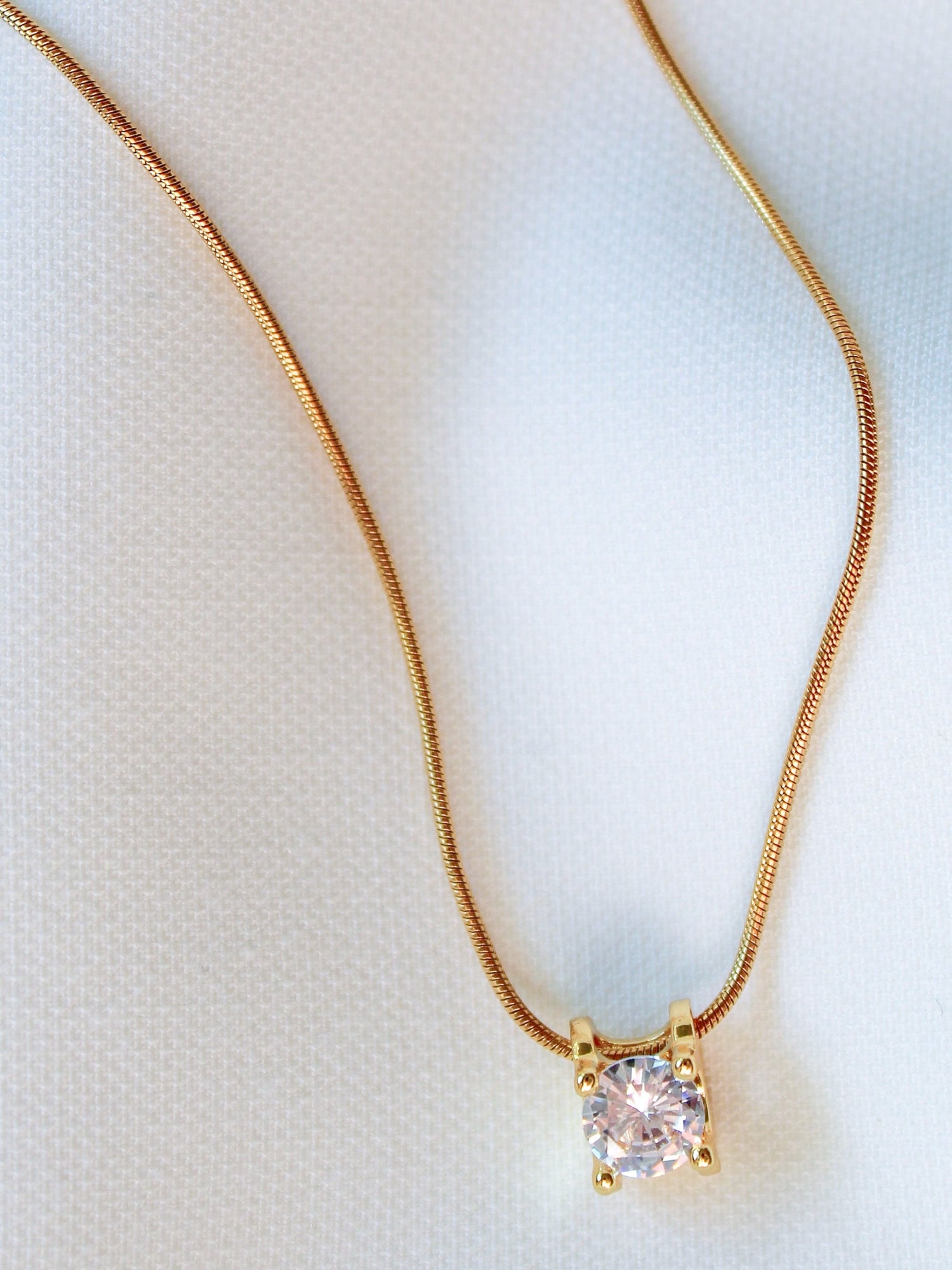 The Jenna Necklace by Kinsey Designs Archives features an 18-inch gold filled chain with a square pendant set with a single sparkling gemstone, displayed on a white fabric background for an elegant touch.