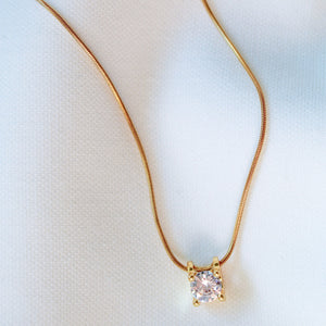 The Jenna Necklace by Kinsey Designs Archives features an 18-inch gold filled chain with a square pendant set with a single sparkling gemstone, displayed on a white fabric background for an elegant touch.