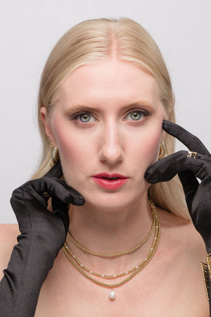 A blonde woman wears black gloves and the Kinsey Designs Kandi Necklace, a gold-filled 16" chain with clasp and extender, gazing at the camera with a neutral expression.