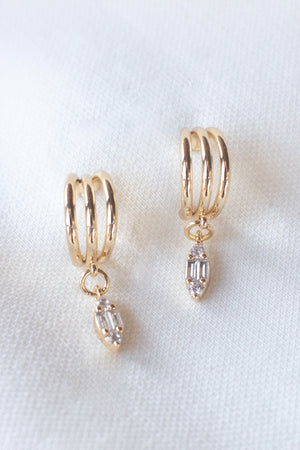 The Keira Drop Hoop earrings by Kinsey Designs Archives feature gold-filled triple hoops with dangling cubic zirconia pendants, displayed on white fabric. Shop these elegant cubic zirconia earrings at our exclusive sample sale.