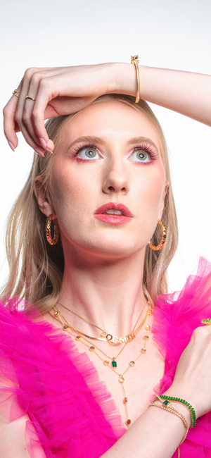 A woman in a pink tulle top wears Kinsey Designs Archives' Kelley Layer, an elegant 18" gold chain, gazing upward with her hand raised above her head.