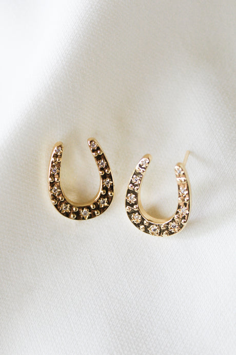 The Kent Earring by Kinsey Designs features gold-filled horseshoe shapes with sparkling gems, shown on a white fabric background. Shop this elegant style now in our sample sale!.