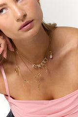 Woman in a pink top wearing layered gold-filled necklaces, featuring the Heart Spacer Slide and Kinsey Charm Bar charms, resting her chin on her hand.