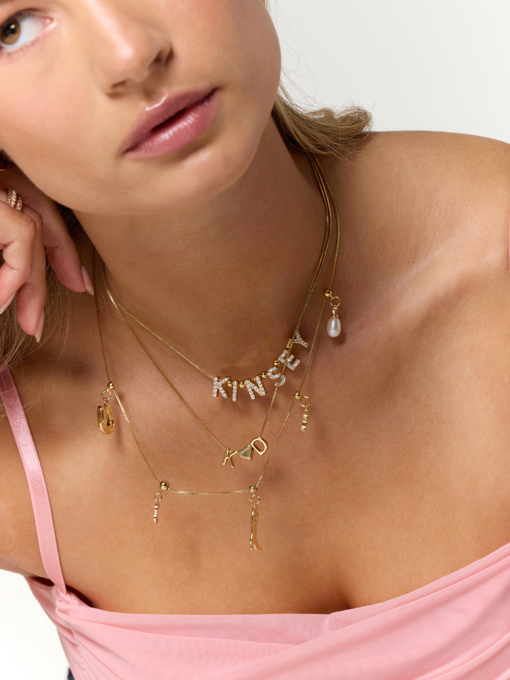 Woman in a pink top wearing layered Kinsey Designs LUX Letter Slide Charms necklaces, styled with gold-filled slide charms and a pearl pendant.
