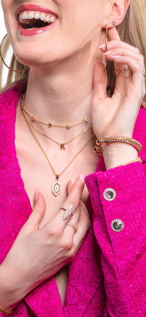 Smiling woman in a pink blazer wearing layered gold jewelry, including rings, bracelets, and the Kinsey Slide Initial Necklace Letter C from the Kinsey Designs Collection.