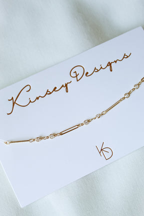 The Leander Necklace by Kinsey Designs, featuring a gold filled chain with small clear stones, is displayed on a white Kinsey Designs card atop a light fabric background. Discover this perfect piece in our sample sale!.