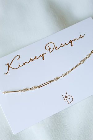 The Leander Necklace by Kinsey Designs, featuring a gold filled chain with small clear stones, is displayed on a white Kinsey Designs card atop a light fabric background. Discover this perfect piece in our sample sale!.