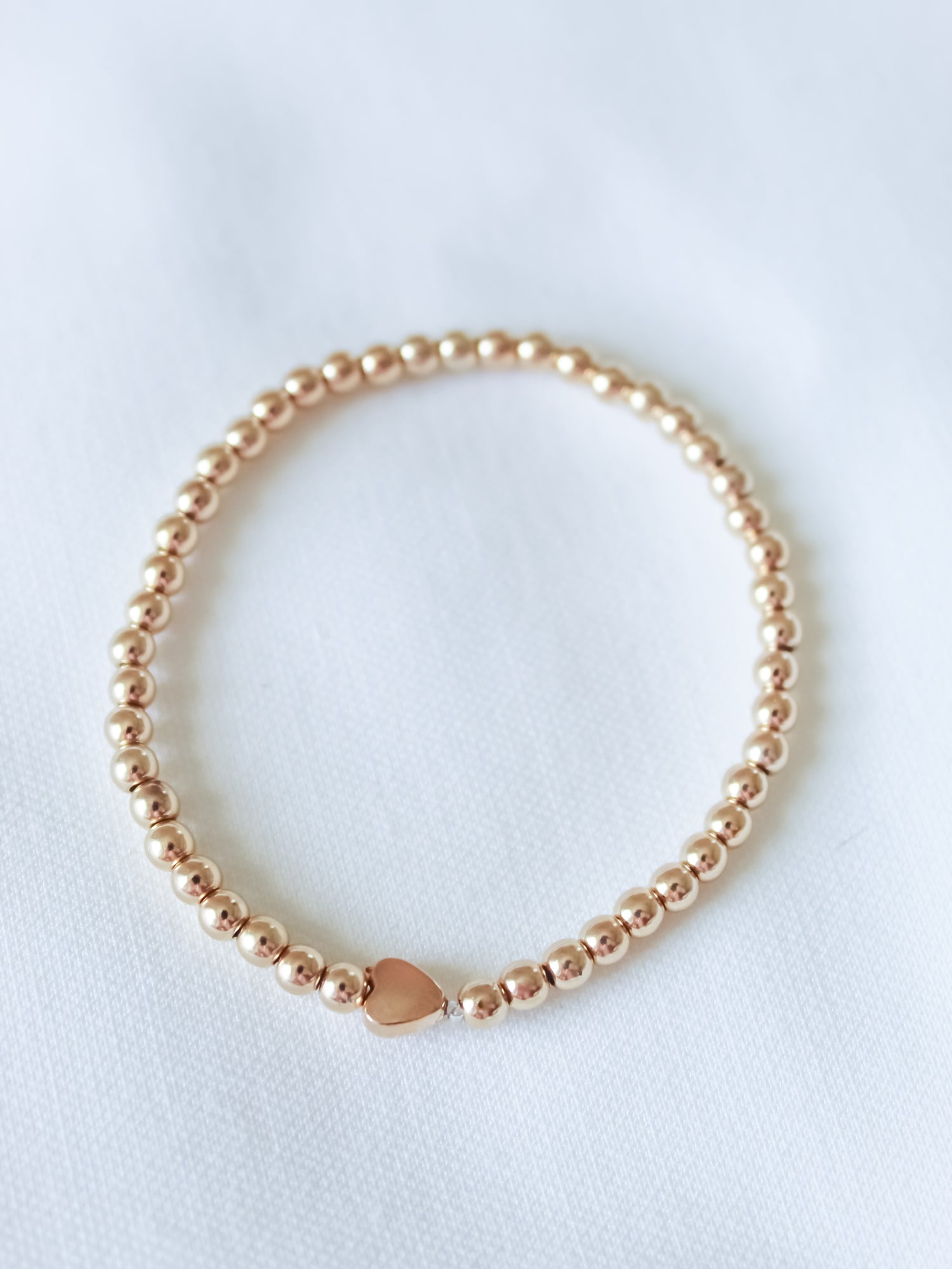 The Liam Heart Bracelet by Kinsey Designs features gold-filled beads and a small heart charm on a white fabric background. With its adjustable stretch design, this bracelet combines elegance and comfort.