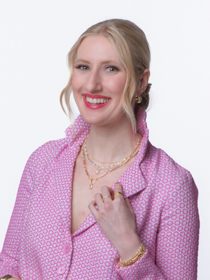 Smiling woman in a pink patterned jacket wears layered gold jewelry, including the elegant Louie Pearl Necklace by Kinsey Designs, against a white background.