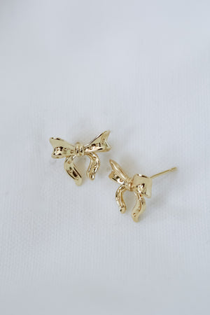 The Kinsey Designs Lova Studs, gold bow earrings displayed on white fabric, add a delicate, feminine touch to any look.