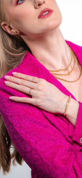 A woman in a textured pink blazer, hand on collar, highlights the Kinsey Designs Lover Necklace—a delicate gold-filled piece—layered with other chains and paired with light pink nail polish.