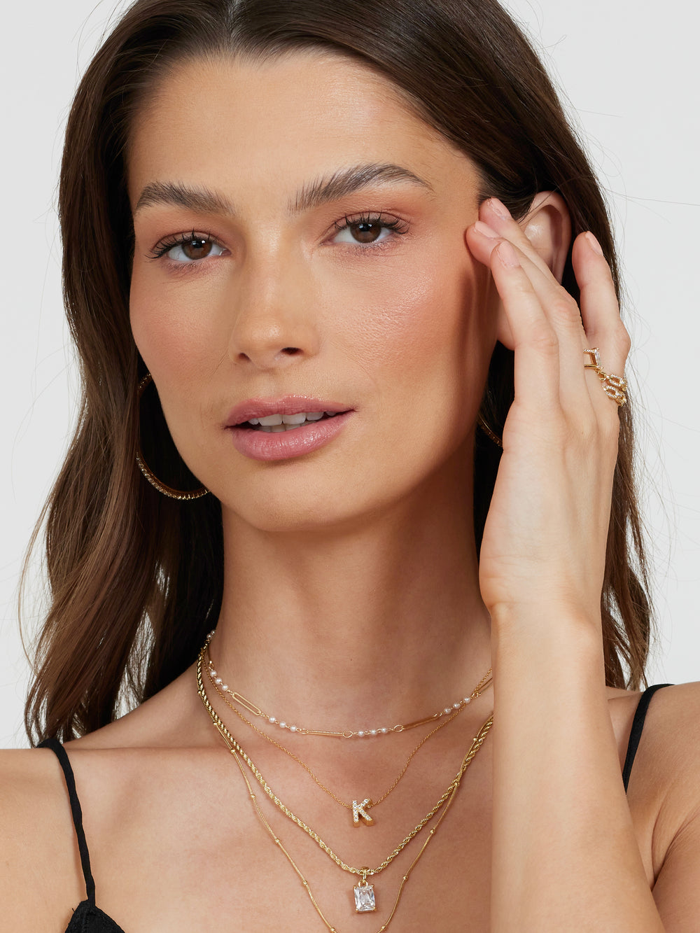 Woman with long brown hair wears the Kinsey Designs Collection LUX Slide Initial Necklace Letter A and hoop earrings, touching her face with one hand.