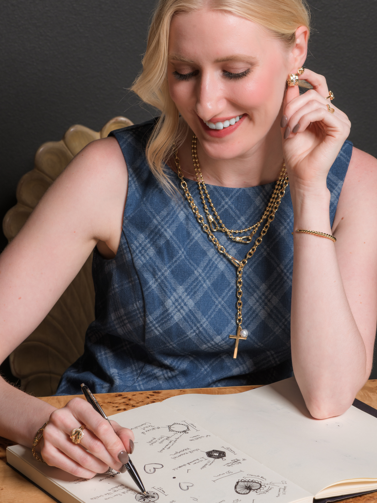 A smiling woman sketches jewelry designs in a notebook while wearing Kinsey Designs’ Marigold Cross Wrap Necklace, adorned with red carnelian beads and a hammered cross pendant, paired with a plaid dress.