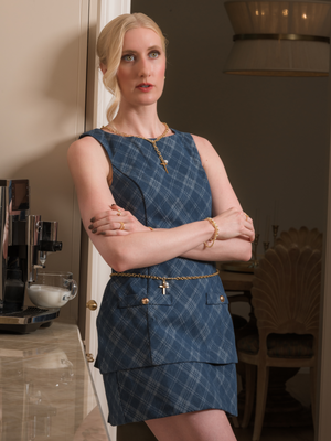 A woman in a blue plaid dress stands with arms crossed, leaning against a counter in a stylish room, wearing the Kinsey Designs Marigold Cross Wrap Necklace.