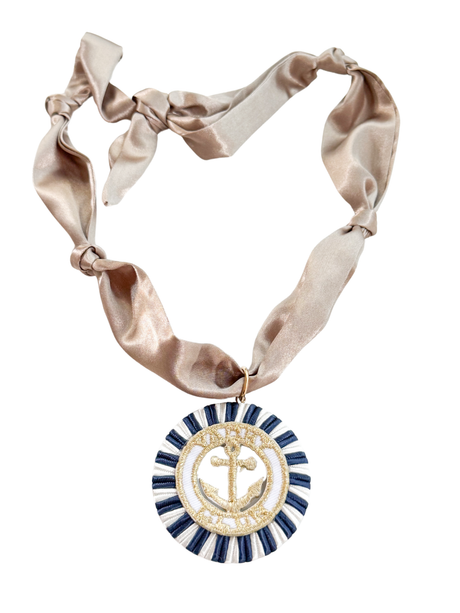 The Matera Scarf Necklace by Kinsey Designs features a gold anchor medallion with blue trim, hanging from a knotted, shiny beige ribbon for a vintage-inspired look.
