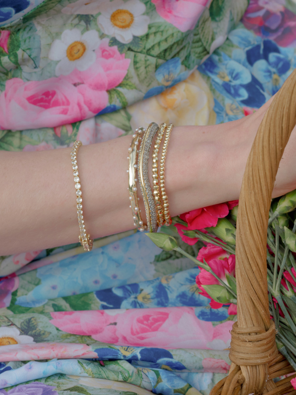 A wrist wearing the Kinsey Designs Multi Faceted Stretch Bead Bracelet holds pink flowers, set against a floral-patterned dress.