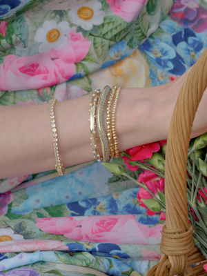 A wrist wearing the Kinsey Designs Multi Faceted Stretch Bead Bracelet holds pink flowers, set against a floral-patterned dress.