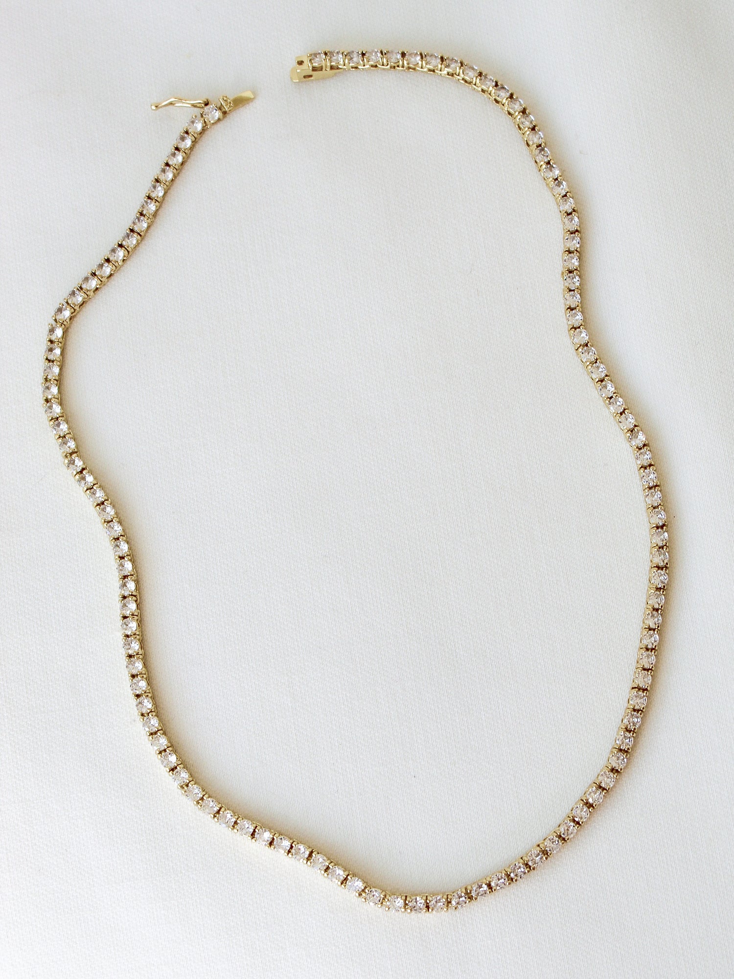 The Malia Tennis Necklace by Kinsey Designs Archives features small, round white stones set in gold fill and is displayed on a white fabric. This elegant 15" necklace suits any occasion.