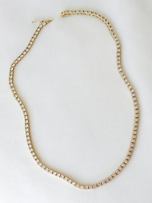 The Malia Tennis Necklace by Kinsey Designs Archives features small, round white stones set in gold fill and is displayed on a white fabric. This elegant 15" necklace suits any occasion.